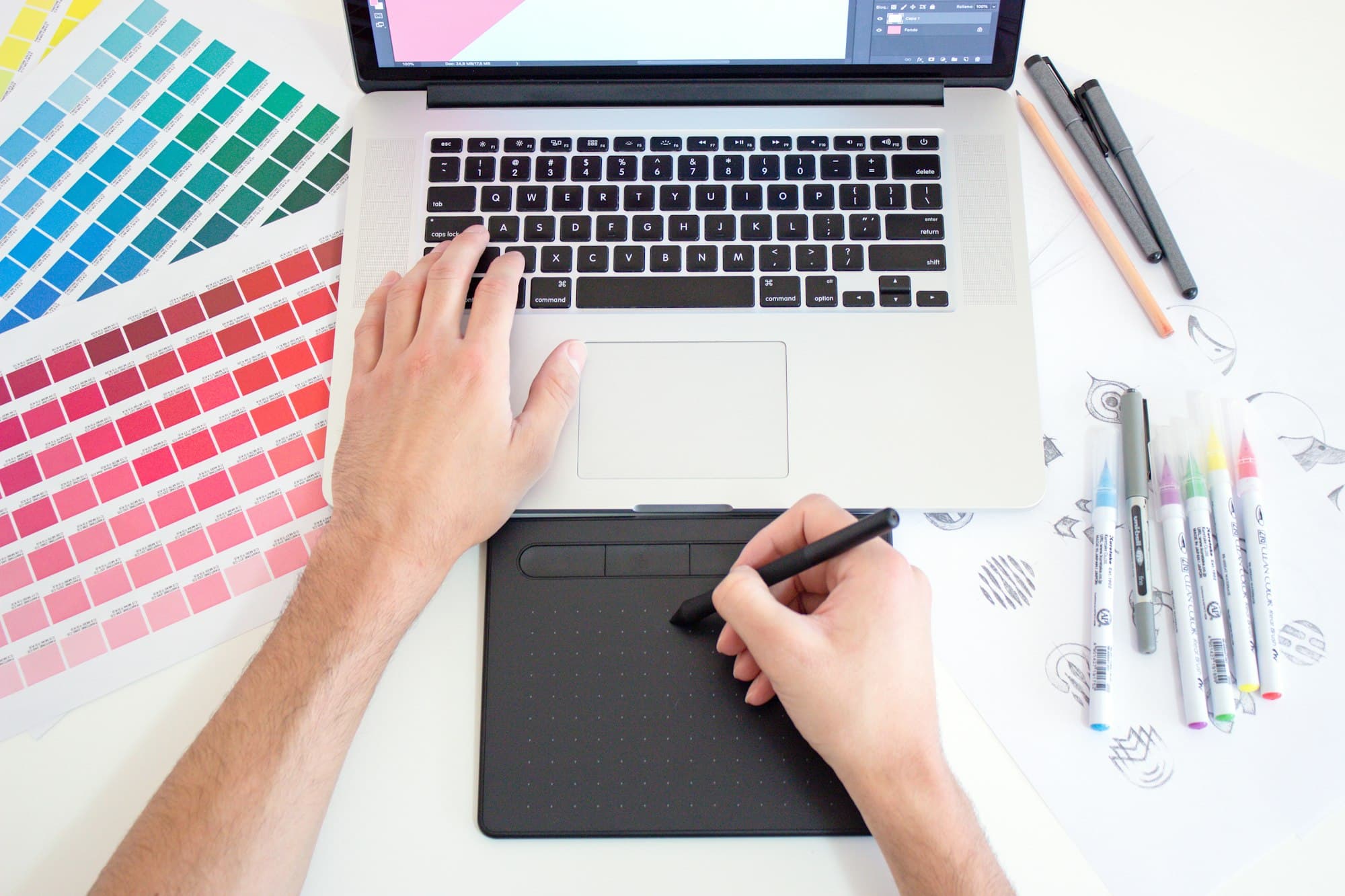 Person arbeitet mit Grafiktablett, Farbkarten und Laptop an einem weißen Tisch